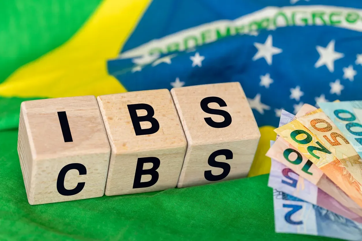 Blocos de madeira com letras “I”, “B” e “S” ao lado de notas de euro sobre a bandeira do Brasil, sugerindo economia e finanças para ibs.