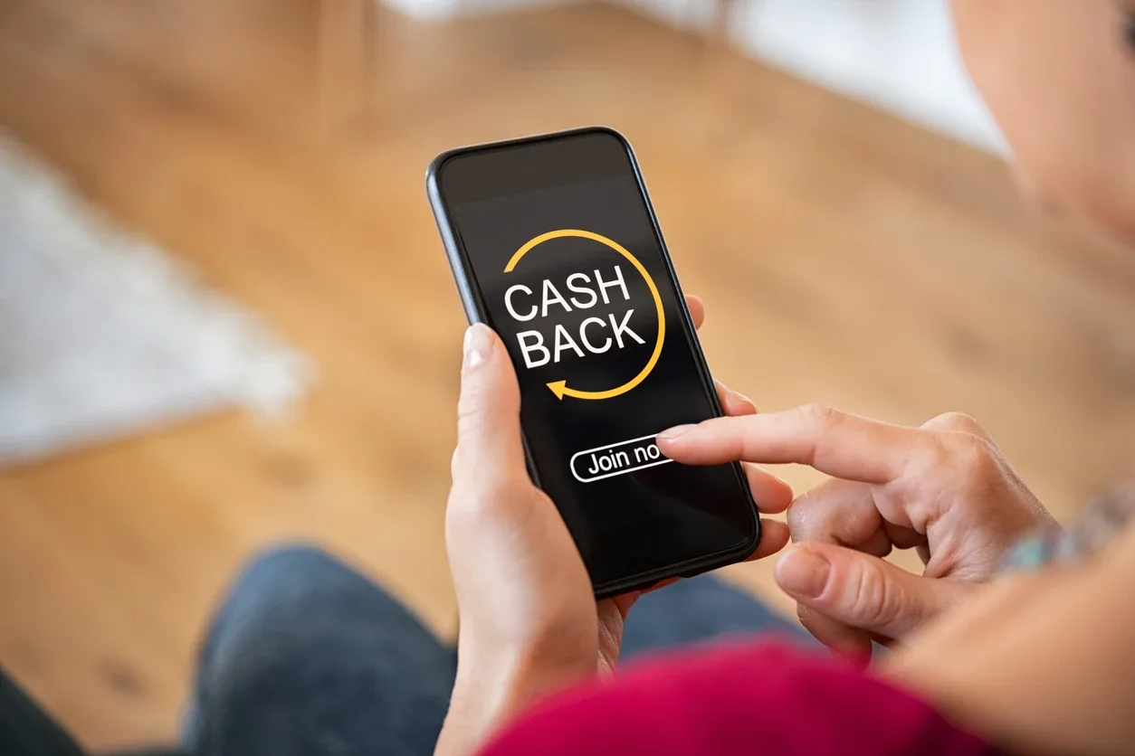 Cashback na reforma tributária: Pessoa segurando um celular com o ícone “CASH BACK” na tela, com mão tocando a opção em um ambiente interno desfocado. Ilustração de cashback.