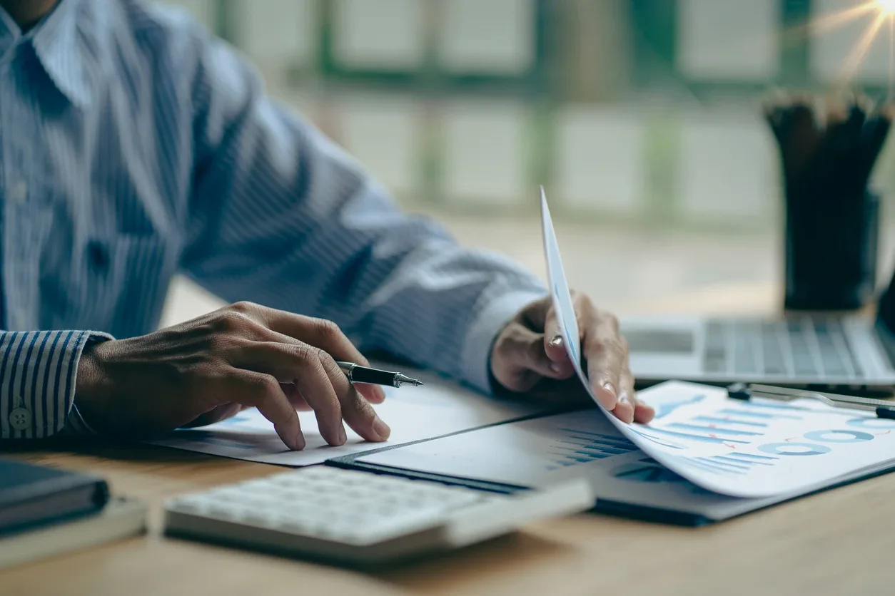 Contas a pagar na reforma tributária: Pessoa em ambiente de escritório analisando papéis e planilhas, com caneta e calculadora sobre a mesa, enquanto registra informações financeiras.