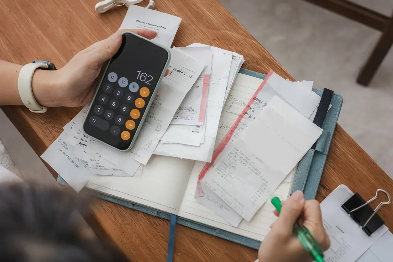 Contas a receber na reforma tributária: Pessoa usando calculadora e mão anotando em documentos e contas sobre uma mesa de escritório com papéis financeiros e conferência de valores