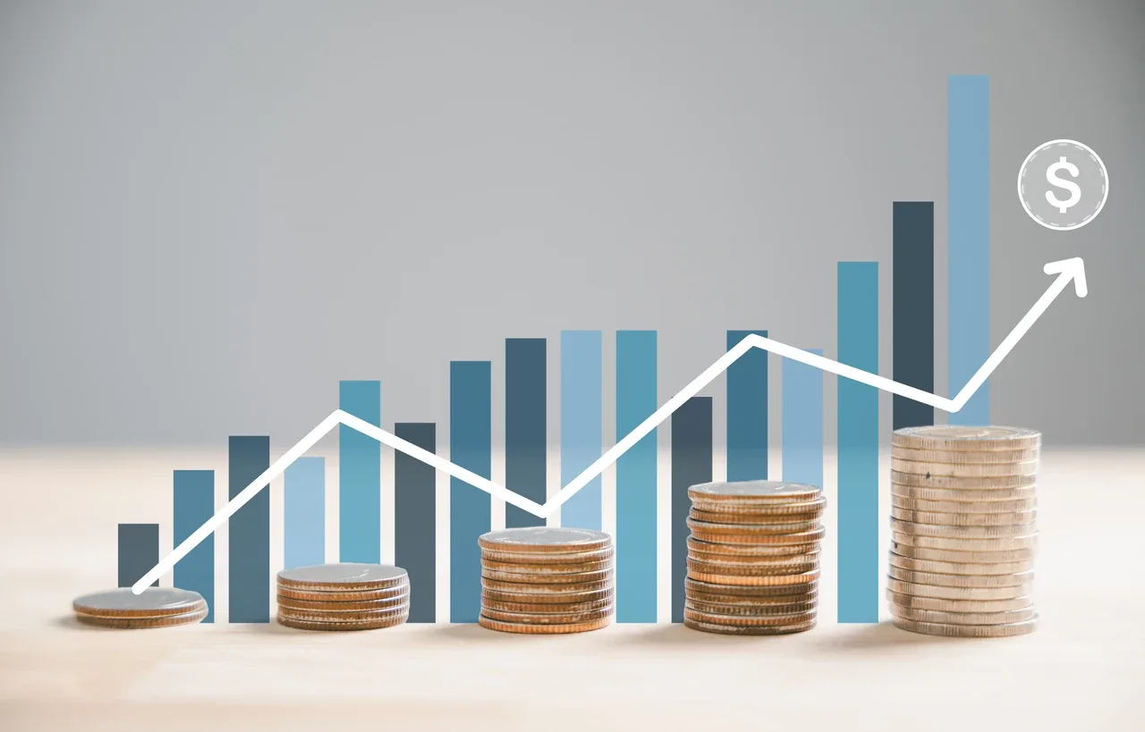 Lucro real e lucro presumido: Gráfico ascendente com linha branca sobre barras azuis e pilhas de moedas sobre mesa, simbolizando crescimento de lucro e rentabilidade.
