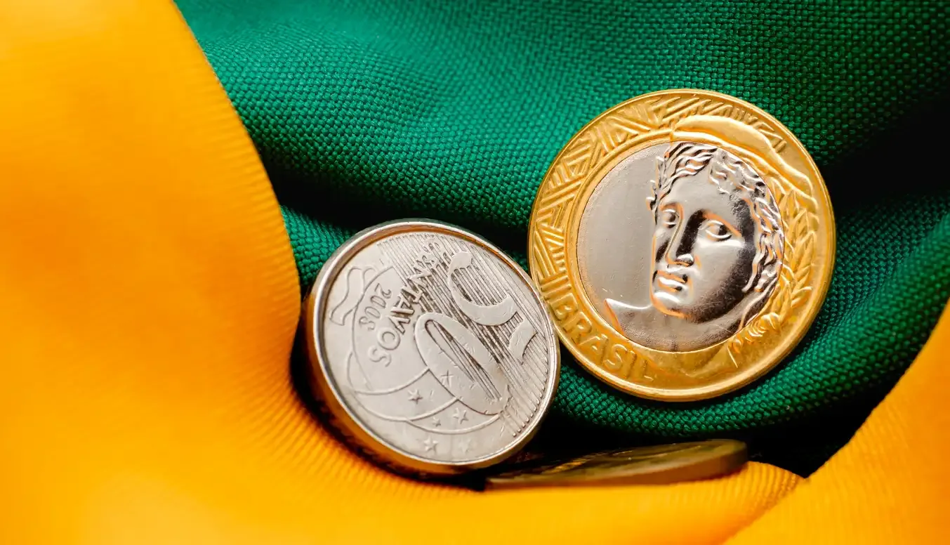 Moedas de ouro e prata sobre tecido verde e amarelo, com destaque para a ideia de finanças e política econômica, sugerindo Reforma tributária.