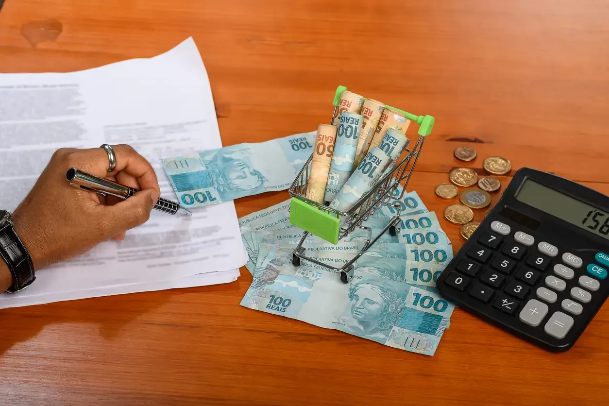 Mesa com documentos, notas de 100 reais, moedas e calculadora digital, representando orçamento base zero e planejamento financeiro