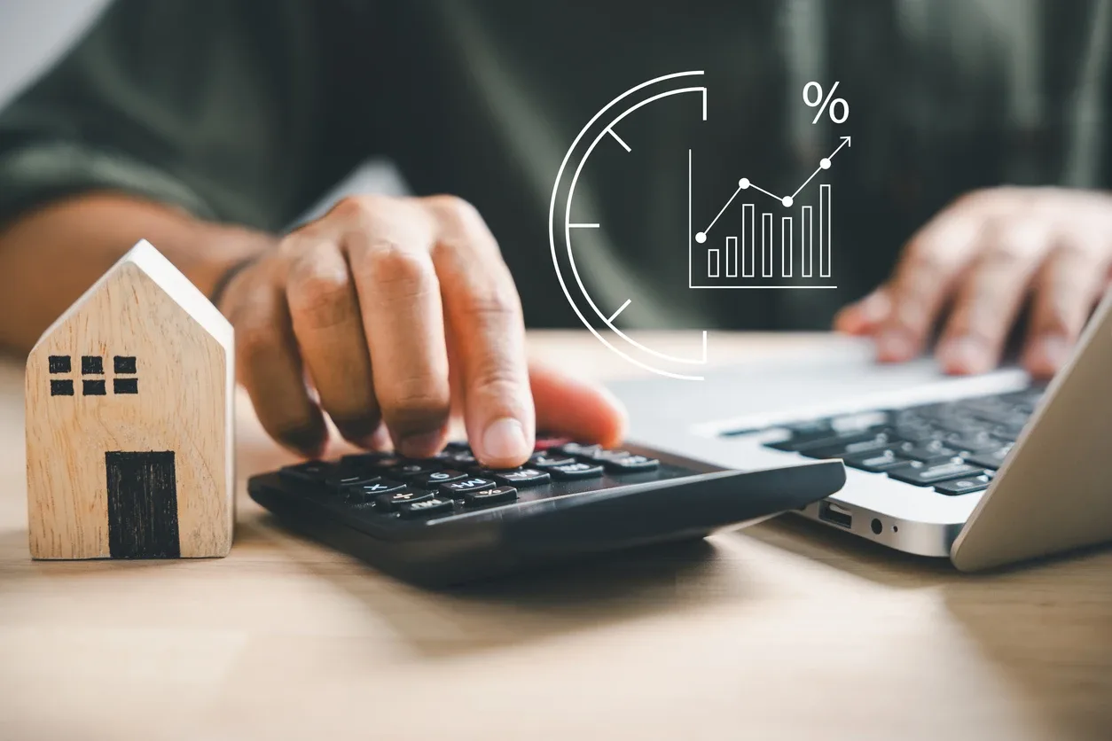 Pessoa calcula valores com calculadora sobre uma mesa, com um modelo de casa ao lado, enquanto um gráfico de porcentagem e ícones de impostos aparecem como referência, sugerindo imposto seletivo.