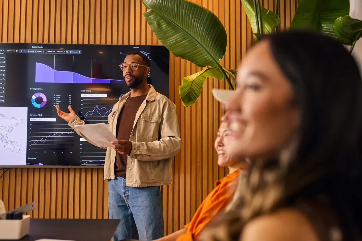 Homem apresentando análises em um monitor com gráficos e indicadores financeiros, enquanto pessoas ao fundo acompanham a reunião em um escritório com plantas. Planejamento financeiro em foco.