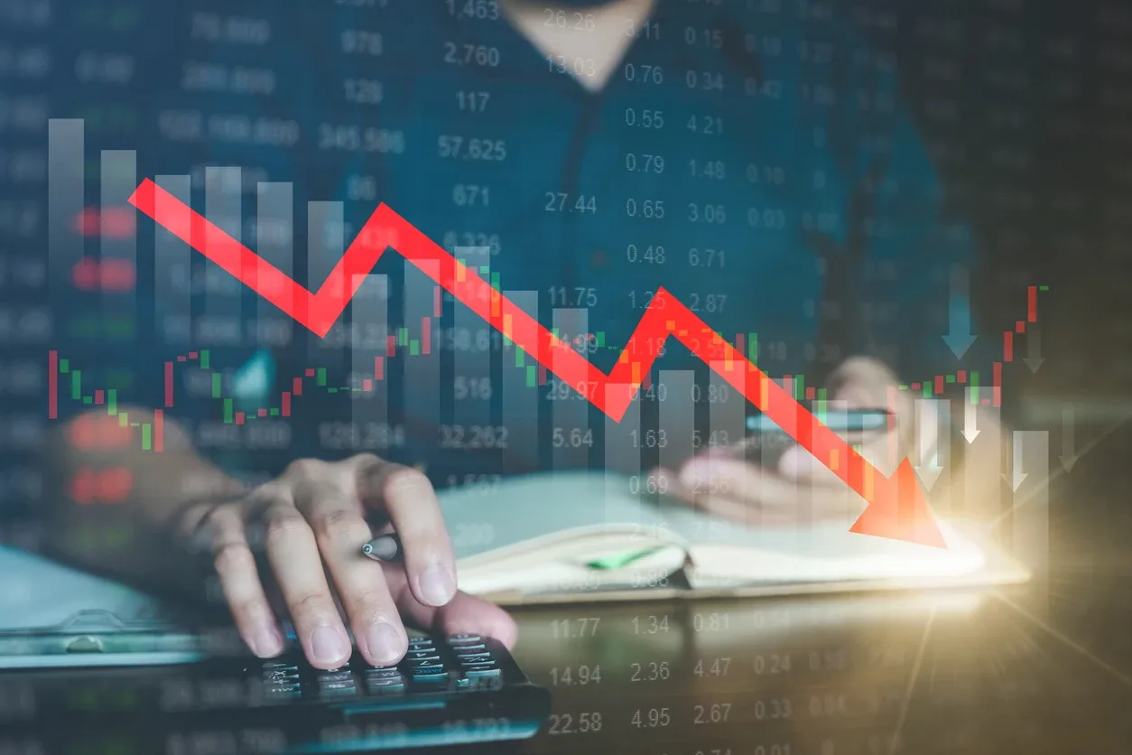 Riscos financeiros representados por um gráfico de queda em vermelho sobre informações de mercado, enquanto uma mão usa uma calculadora com números e barras ao fundo.