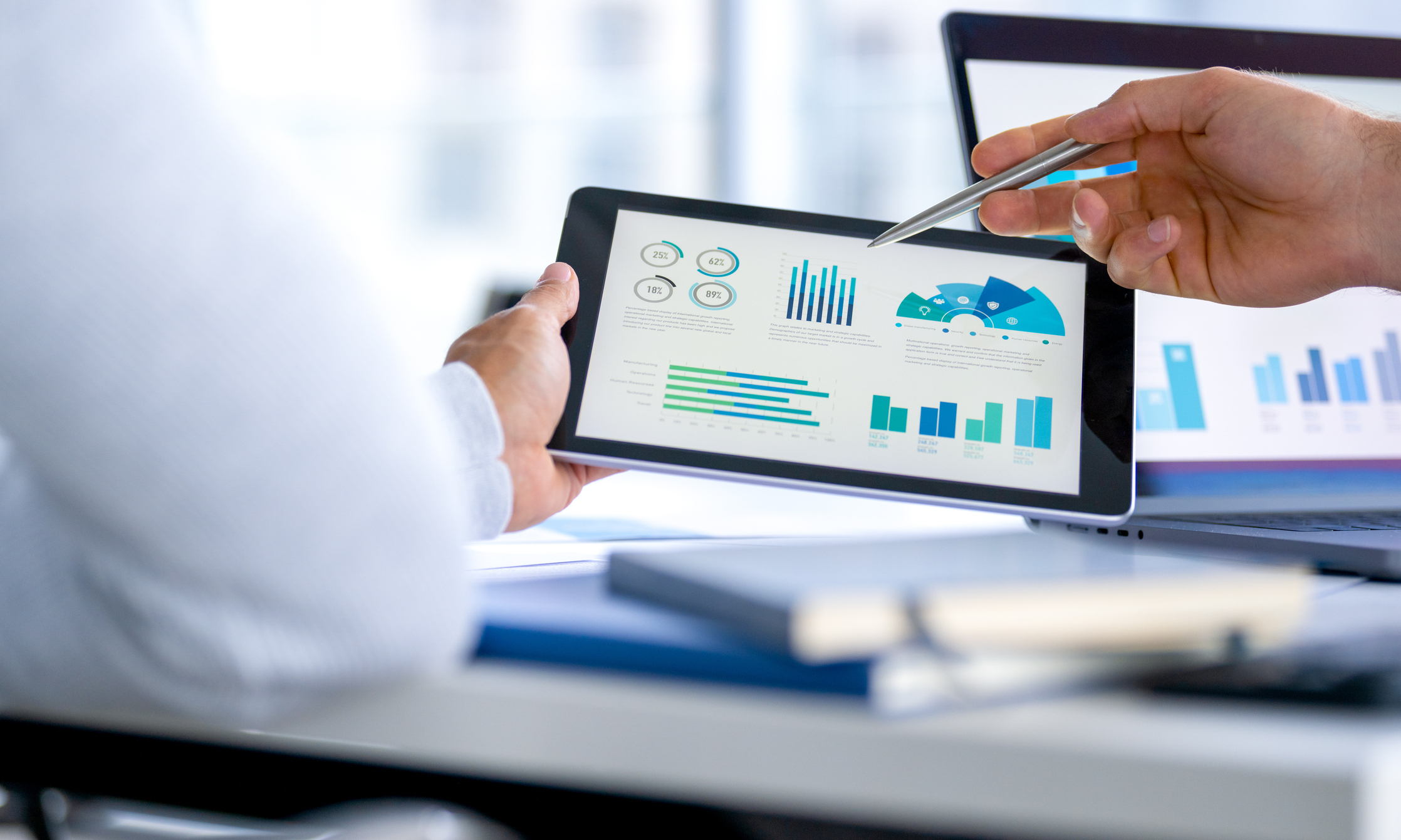 Dois empresários se reúnem e analisam um orçamento empresarial com gráficos e tabelas em um tablet digital e em um notebook.
