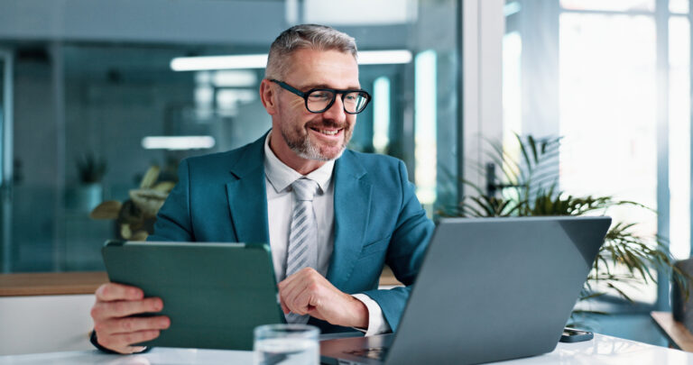 Homem de negócios sorridente usando tablet e laptop em escritório moderno, representando gestão de cobrança eficiente e profissional.