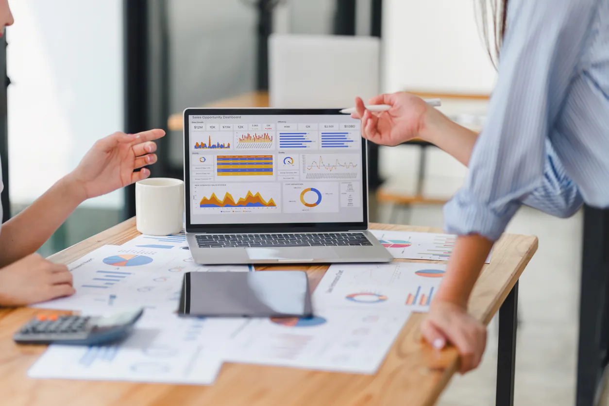 Duas pessoas analisando gráficos de gestão financeira para agências em uma reunião de negócios com laptop, papéis e gráficos de desempenho.