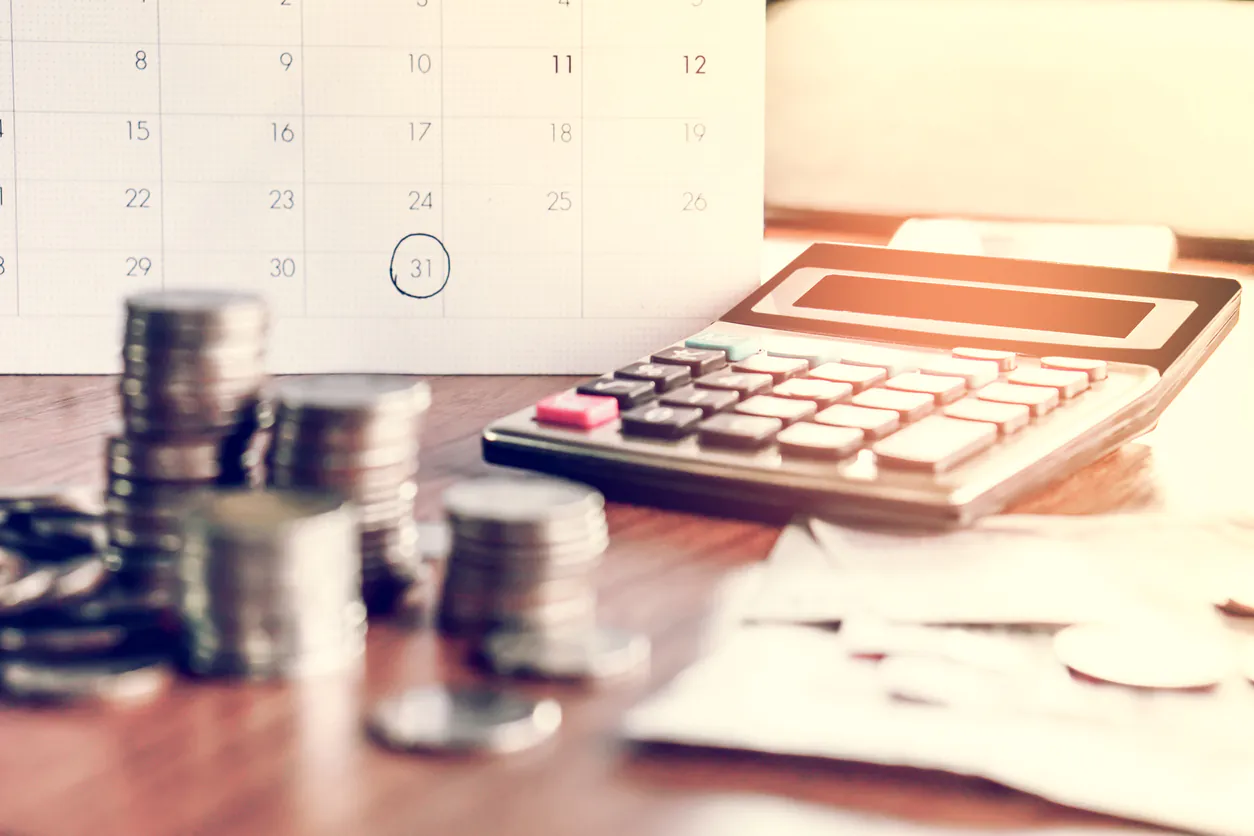 Imagem de uma calculadora financeira ao lado de pilhas de moedas, uma agenda marcada e papéis, simbolizando gestão financeira e o prazo médio de recebimento.