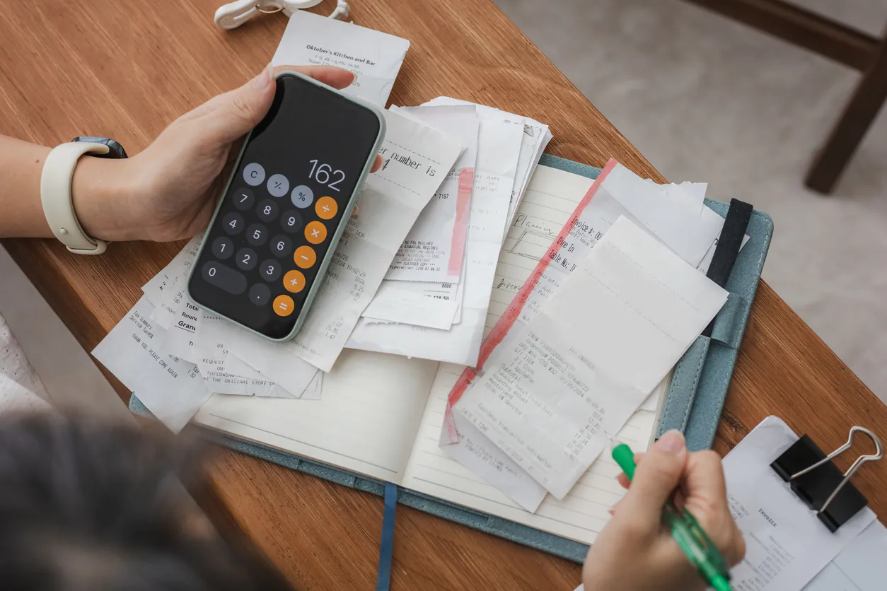 Pessoa usando smartphone e anotando em caderno, com pilhas de contas e papel na mesa, representando automação contas a pagar comparativo de soluções.