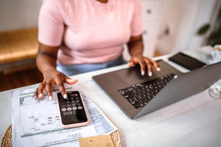 Pessoa gerenciando controle de contas a receber com planilhas, smartphone e computador portátil em ambiente de escritório moderno.