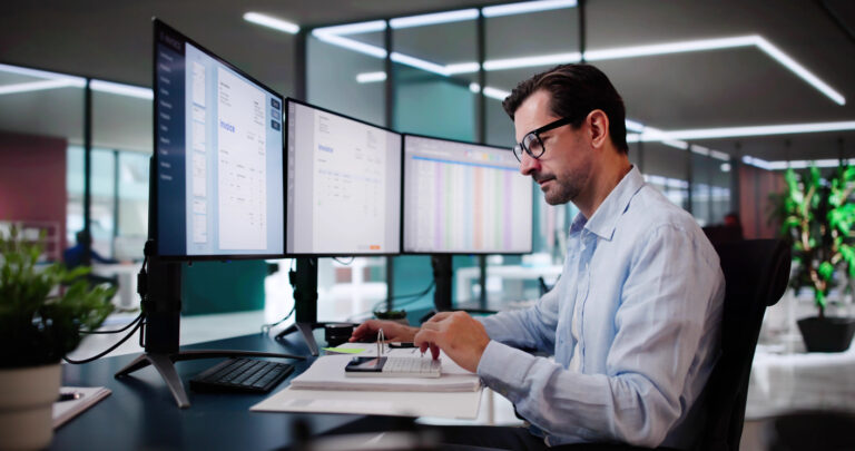 Homem profissional trabalha com múltiplos monitores em um escritório moderno, analisando dados e gráficos em ambiente de alta tecnologia.