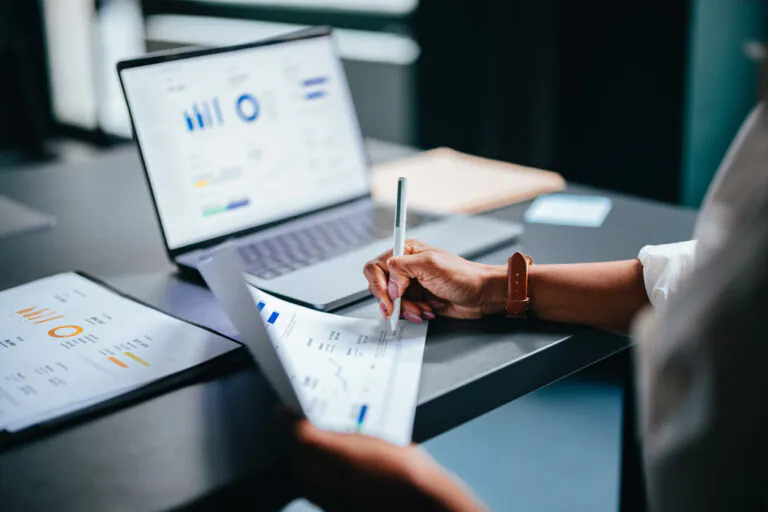 Apropriação de despesas em ambiente corporativo: mãos analisam relatórios e gráficos no notebook enquanto alguém registra informações com caneta sobre documentos financeiros na mesa