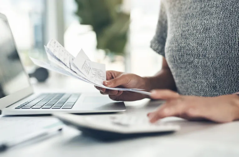 Pessoa usando um laptop enquanto segura um documento de comprovante de despesa com dados impressos em papel, sugerindo conferência ou organização de gastos.