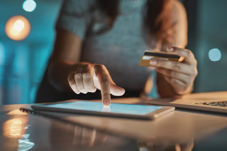 Pessoa em ambiente de escritório aponta para um tablet enquanto segura um cartão de crédito e usa um computador, sugerindo conciliação de cartão e conferência de transações financeiras.