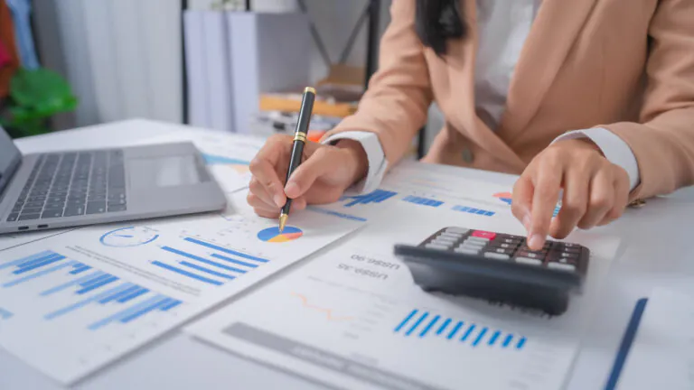 Controle de despesas corporativas: profissional analisando planilhas e gráficos financeiros em uma mesa com calculadora e laptop, enquanto registra dados com caneta.