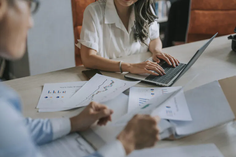 Equipe em reunião corporativa analisando relatórios e planilhas no computador, com gráficos em documentos sobre Despesas administrativas e gestão financeira no escritório