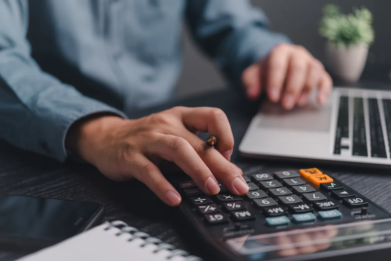 Pessoa analisando despesas financeiras no escritório, usando calculadora e computador, com anotações em caderno sobre a mesa e mão apontando números na tela.