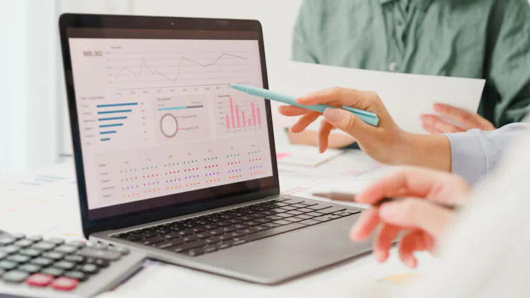 Pessoa analisando gráficos e indicadores de Expense management em um notebook, apontando com caneta sobre relatórios financeiros na tela, enquanto planeja despesas na mesa.