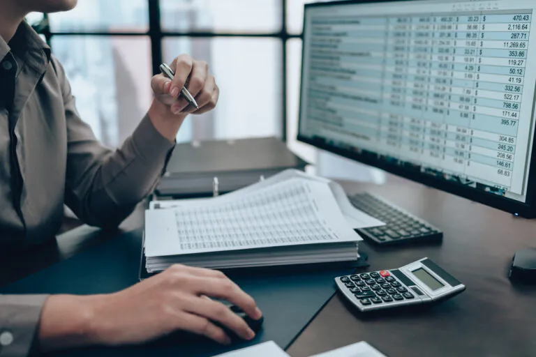 Pessoa em ambiente corporativo analisando documentos e números em um computador, enquanto calcula valores com calculadora e caneta sobre a prestação de contas de cartão corporativo.