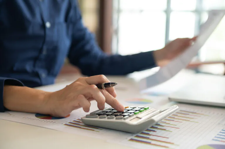 Pessoas analisando planilhas e usando uma calculadora em escritório, com anotações ao fundo, para apoiar cálculos contábeis como depreciação e amortização.