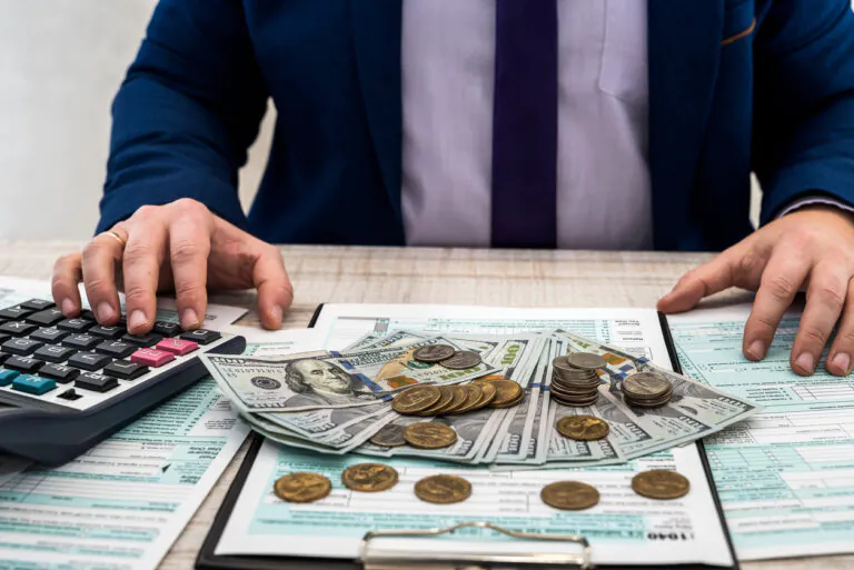 Pessoa em ambiente de escritório calculando contas com calculadora e analisando cédulas e moedas sobre documentos financeiros, tema de despesas dedutiveis.
