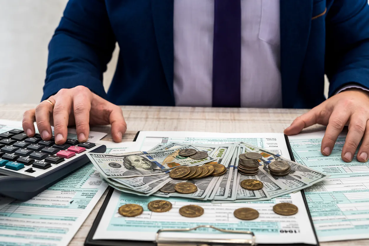 Pessoa em ambiente de escritório calculando contas com calculadora e analisando cédulas e moedas sobre documentos financeiros, tema de despesas dedutiveis.