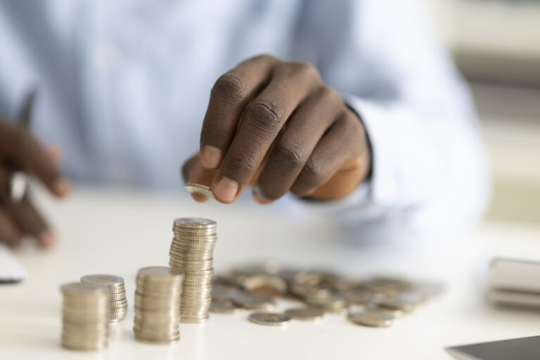 Mão colocando uma moeda sobre uma pilha de moedas, enquanto outras pessoas analisam contas e dinheiro sobre uma mesa, sugerindo controle e planejamento de despesas fixas e variáveis.