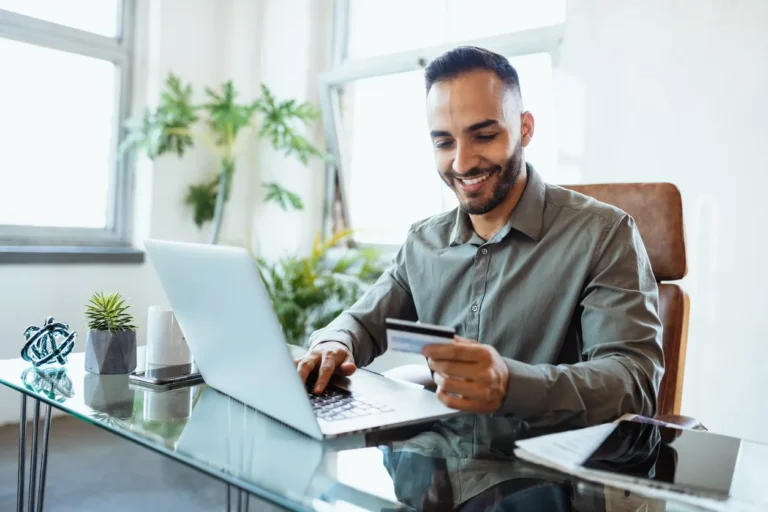 Homem sorridente em escritório moderno usa notebook e segura um cartão na mão enquanto trabalha, conceito de melhores cartões corporativos para pagamentos e compras