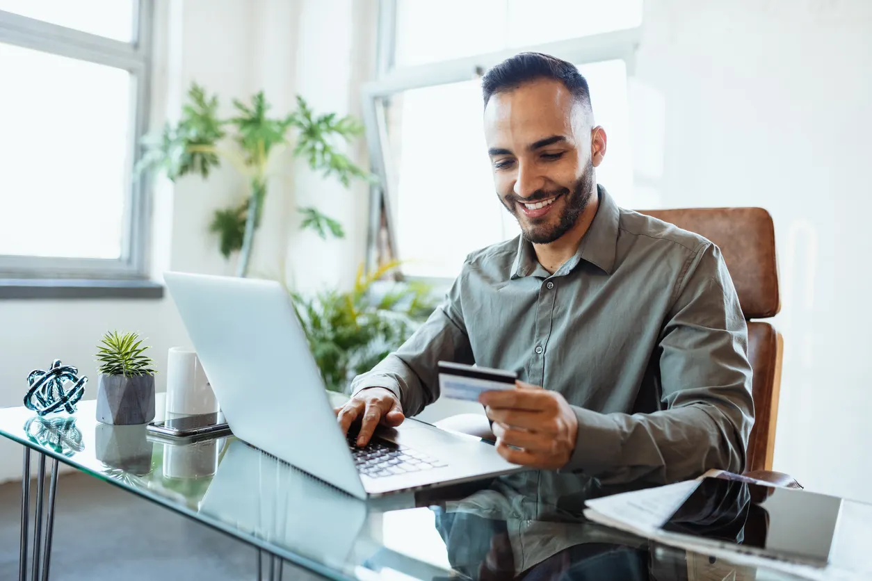 Homem sorridente em escritório moderno usa notebook e segura um cartão na mão enquanto trabalha, conceito de melhores cartões corporativos para pagamentos e compras